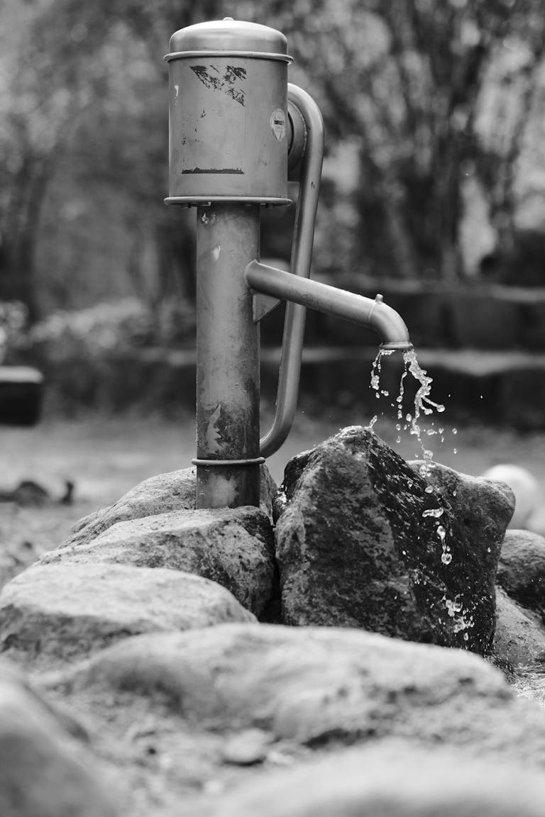 Black and white image of a rustic outdoor water pump with water flowing over stones.
